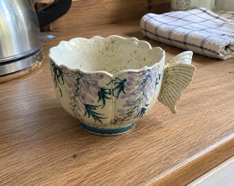 May include: A ceramic mug with a scalloped rim and a handle shaped like a butterfly wing. The exterior features a hand-painted pattern of purple wisteria flowers and green leaves on a cream-colored speckled base with a blue ring at the bottom.