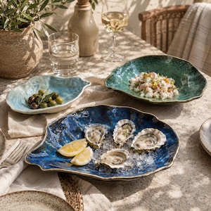 May include: Three irregular, shell-shaped ceramic bowls in shades of light blue, deep green, and dark blue. The dark blue bowl contains four raw oysters on ice with lemon wedges, the green bowl holds a seafood salad, and the light blue bowl holds olives.