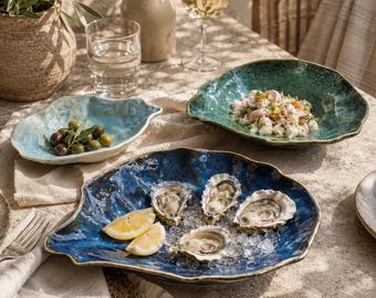 May include: Three irregular, shell-shaped ceramic bowls in shades of light blue, deep green, and dark blue. The dark blue bowl contains four raw oysters on ice with lemon wedges, the green bowl holds a seafood salad, and the light blue bowl holds olives.