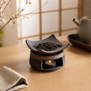Puede incluir: Un calentador de t&eacute; de cer&aacute;mica marr&oacute;n oscuro con una placa superior cuadrada y texturizada que contiene t&eacute; a granel. La base presenta una abertura circular con una peque&ntilde;a vela de t&eacute; encendida, mostrando un acabado r&uacute;stico y moteado.