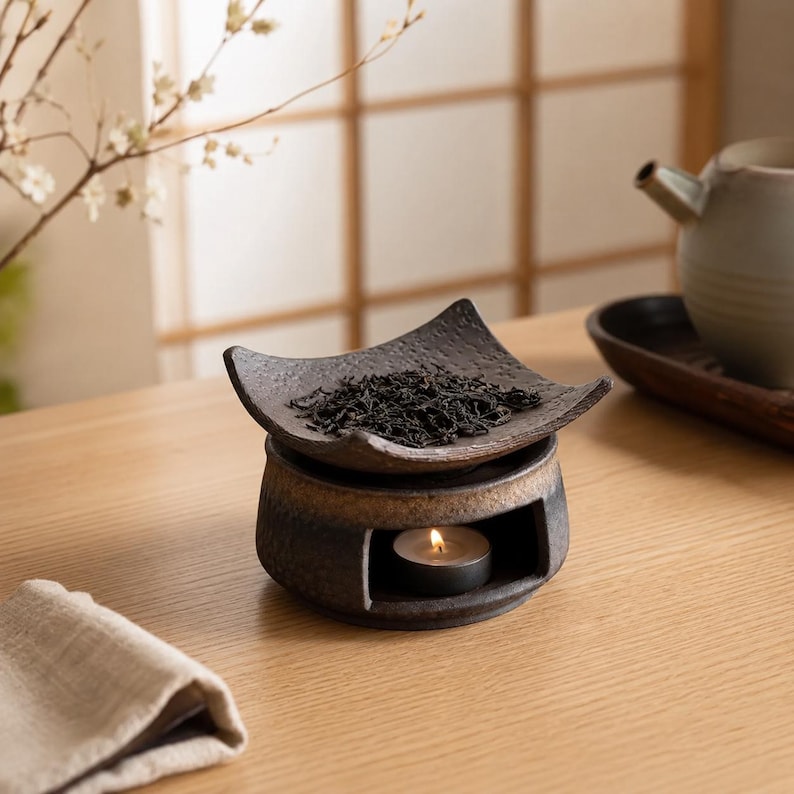 Puede incluir: Un calentador de t&eacute; de cer&aacute;mica marr&oacute;n oscuro con una placa superior cuadrada y texturizada que contiene t&eacute; a granel. La base presenta una abertura circular con una peque&ntilde;a vela de t&eacute; encendida, mostrando un acabado r&uacute;stico y moteado.