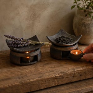Puede incluir: Dos quemadores de aceite de cer&aacute;mica oscura y texturizada con cuencos cuadrados de bordes curvos. Uno contiene ramas de lavanda morada seca y el otro hojas de t&eacute; oscuras. Cada base hueca alberga una vela de t&eacute; encendida.