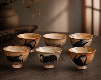 May include: Six ceramic teacups with a rustic, hand-painted design. Each cup features a black cat illustration against a warm, earthy background. The cups are arranged on a dark wooden surface, showcasing their unique, artistic style.