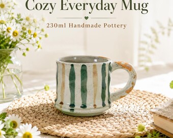 May include: A ceramic mug with a speckled off-white glaze featuring vertical green and tan stripes. The text reads Cozy Everyday Mug 230ml Handmade Pottery. The mug has a thick handle with orange speckles and a rounded base.