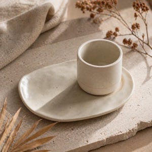 Könnte beinhalten: Ein beiges Keramik-Tassen- und Untertassen-Set. Die zylindrische Tasse steht auf einer ovalen Untertasse. Das Set steht auf einer strukturierten Oberfläche, was eine minimalistische Ästhetik erzeugt.