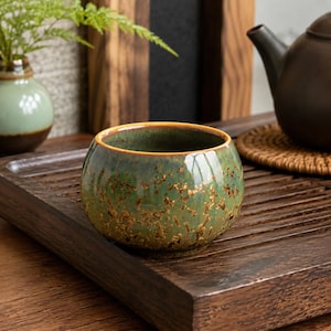 May include: A small, round ceramic tea cup featuring a mottled green glaze with golden-brown flecks and a smooth, glossy finish. The cup has a slightly tapered rim and a rounded body, showcasing a reactive glaze pattern that creates a unique texture.