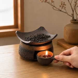Puede incluir: Un calentador de t&eacute; de cer&aacute;mica marr&oacute;n oscuro con una placa superior cuadrada y texturizada que contiene hojas de t&eacute; sueltas. La base tiene una abertura para una peque&ntilde;a taza redonda con asa que contiene una vela encendida con llama.