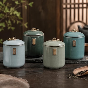 May include: Four ceramic tea canisters in shades of blue and green. Each canister has a lid with a brass ring handle and a small rectangular label. The canisters are cylindrical and sit on a dark wooden surface.
