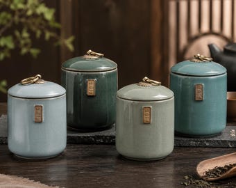 Puede incluir: Cuatro botes de té de cerámica en tonos azules y verdes. Cada bote tiene una tapa con un asa de anillo de latón y una pequeña etiqueta rectangular. Los botes son cilíndricos y se asientan sobre una superficie de madera oscura.