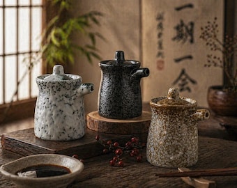 May include: Three ceramic sauce dispensers with lids. One is white with black speckles, another is black, and the third is brown with speckles. They are on a wooden surface, with a small bowl of sauce in the foreground.