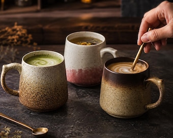 May include: Three ceramic mugs with speckled textures and gradient color finishes. One mug contains green matcha, one holds a light herbal tea, and the third contains coffee with latte art, being stirred by a gold-toned metal spoon.