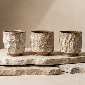 May include: Three ceramic cups with a speckled beige and brown finish. The first cup has a faceted geometric texture, the second features a grid of incised lines, and the third displays vertical wavy grooves. Each cup has a distinct base and rim.