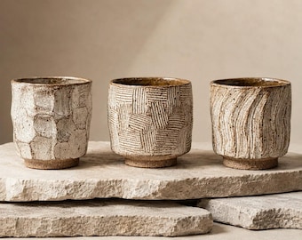 Puede incluir: Tres tazas de cerámica con un acabado moteado en beige y marrón. La primera taza tiene una textura geométrica facetada, la segunda presenta una cuadrícula de líneas incisas y la tercera muestra surcos ondulados verticales.