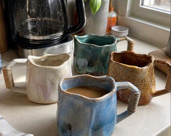 May include: Four ceramic mugs with irregular, wavy rims and handles wrapped in light-colored rope. The mugs are finished in glossy glazes of cream, teal, brown, and blue. One blue mug is filled with coffee, and all feature a textured, organic shape.
