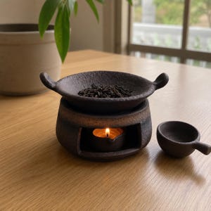 Puede incluir: Un quemador de aceite de cer&aacute;mica marr&oacute;n oscuro con acabado texturizado y dos peque&ntilde;as asas laterales. El quemador cuenta con una abertura central que contiene una vela de t&eacute; encendida, coronada por un cuenco poco profundo con hojas de t&eacute; sueltas y una peque&ntilde;a cuchara a juego.