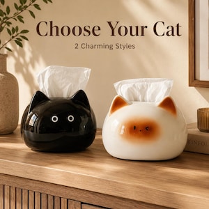 May include: Two cat-shaped tissue holders with white tissues emerging from the top. One is glossy black with white circular eyes, and the other is cream-coloured with brown ear tips and a brown face patch. Text reads: Choose Your Cat 2 Charming Styles.