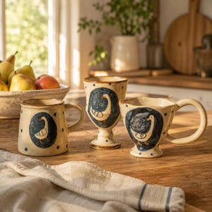 Op de afbeelding: Drie crèmekleurige keramische vaten met een gespikkelde afwerking en donkerblauwe stippen. Elk stuk is voorzien van een handgeschilderde illustratie van een gans in een donkerblauw ovaal kader. De set bevat een mok en twee kelkvormige bekers.