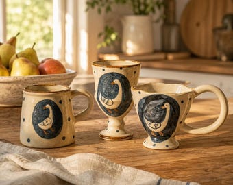 May include: Three cream-colored ceramic vessels with a speckled finish and dark blue polka dots. Each piece features a hand-painted illustration of a goose inside a dark blue oval frame. The set includes a mug, a stemmed goblet, and a wide-rimmed cup.