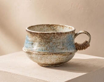 May include: A handcrafted ceramic mug with a unique design. The mug features a light blue and brown color scheme with a textured surface. The handle is wrapped with a natural rope. This mug is perfect for enjoying your favorite hot beverages.