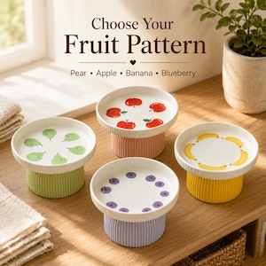 Puede incluir: Cuatro cuencos de cerámica con pedestal y motivos frutales. El texto dice Choose Your Fruit Pattern, Pear, Apple, Banana, Blueberry. Cada cuenco tiene una base de color único: verde, rosa, amarillo y morado claro, con ilustraciones de frutas.