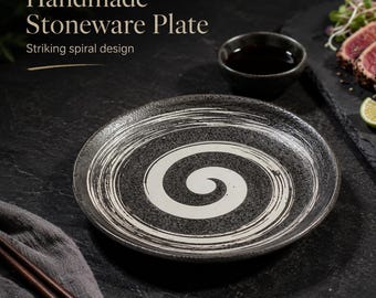 May include: A round stoneware plate featuring a black textured surface with a prominent white painted spiral design. The plate is presented alongside a small black dipping bowl, a pair of wooden chopsticks, and a slate board with food items.