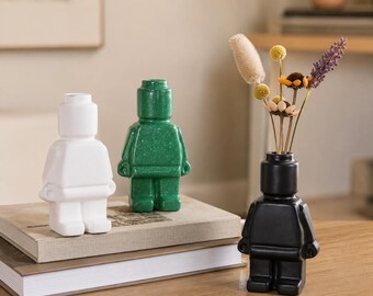 May include: Three ceramic figurines shaped like toy blocks. One is white, one is green, and the third is black and holds dried flowers. The figurines are on a wooden surface with books.