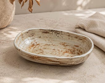 May include: An oval ceramic bowl with a cream-colored glaze and a distressed, rustic finish featuring visible brown brushstrokes throughout the interior and exterior surfaces, creating a textured appearance with an irregular, organic rim shape.