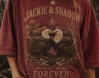May include: A maroon oversized t-shirt featuring a graphic print of two bald eagles facing each other. The text reads JACKIE & SHADOW at the top and TWO HEARTS ONE SKY FOREVER at the bottom. The design includes a compass star and pine trees.