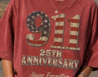 May include: A red, short-sleeved t-shirt featuring a graphic with the number 911 filled with an American flag pattern. Below the number, the text reads 25TH ANNIVERSARY and Never Forgotten, accompanied by three small stars in a horizontal row.