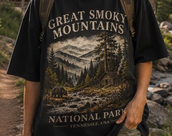 May include: Black t-shirt with a graphic of the Great Smoky Mountains National Park. The design features a mountain landscape, a cabin, and the text "Great Smoky Mountains National Park, Tennessee, USA". The shirt is worn outdoors.