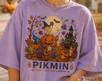 May include: A light purple short-sleeve t-shirt featuring a colorful graphic of Pikmin characters, pumpkins, and a haunted house. The text reads PIKMIN PICK, PLANT, AND SPOOK! The design includes autumn leaves, a full moon, and bats in a fall setting.