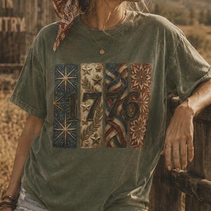 May include: Olive green t-shirt with a patriotic 1776 design. The design features stars, stripes, and floral patterns in red, white, and blue. The shirt is worn with denim shorts and a bandana.