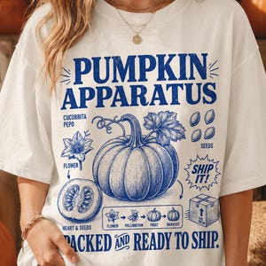 May include: A white short-sleeve t-shirt featuring a blue graphic print with the text PUMPKIN APPARATUS, SHIP IT, and PACKED AND READY TO SHIP. The design includes illustrations of a pumpkin, a cross-section, seeds, a flower, and a box.