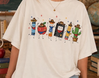 May include: An off-white short-sleeve t-shirt featuring a graphic print of six anthropomorphic school supplies wearing cowboy hats and bandanas, including a crayon, apple, glue stick, notebook, pencil, and cactus, arranged in a horizontal row.