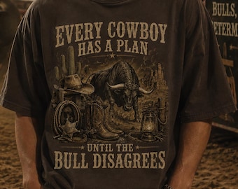 May include: A dark charcoal gray short-sleeve t-shirt featuring a graphic print of a bull, a cowboy hat, boots, a lasso, and a lantern. The text reads: EVERY COWBOY HAS A PLAN UNTIL THE BULL DISAGREES. The shirt has a crew neckline and a relaxed fit.