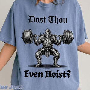 Op de afbeelding: Lichtblauw T-shirt met de tekst "Dost Thou Even Hoist?" boven en onder een afbeelding van een ridder die een halter optilt. De ridder draagt een volledig harnas en de halter heeft decoratieve uiteinden.