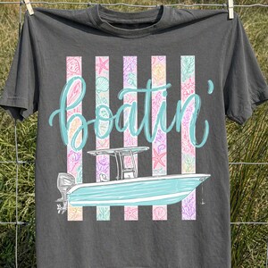 May include: Dark gray t-shirt with a boat graphic and the word "boatin'" in turquoise script. The boat is light blue, and the background features vertical stripes with seashell and starfish patterns. The shirt is hanging on a clothesline.
