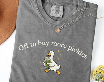 May include: A gray t-shirt with the phrase "Off to buy more pickles" in a curved white font. A cartoon duck carrying a jar of pickles is in the center. A gold necklace is draped across the shirt.