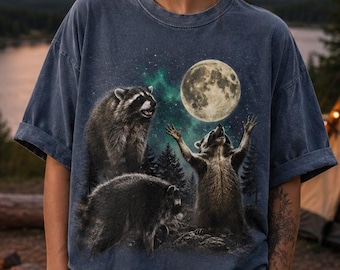May include: A slate blue short-sleeve t-shirt featuring a graphic print of three raccoons in a forest setting at night. One raccoon stands with arms raised toward a large, bright full moon in a starry sky, while two others are nearby.
