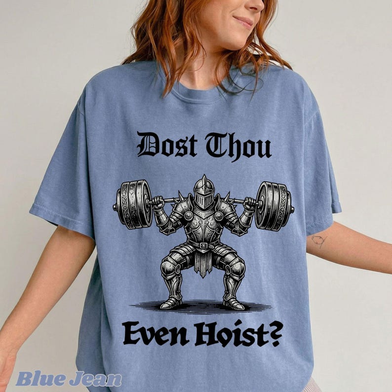 Op de afbeelding: Lichtblauw T-shirt met de tekst "Dost Thou Even Hoist?" boven en onder een afbeelding van een ridder die een halter optilt. De ridder draagt een volledig harnas en de halter heeft decoratieve uiteinden.