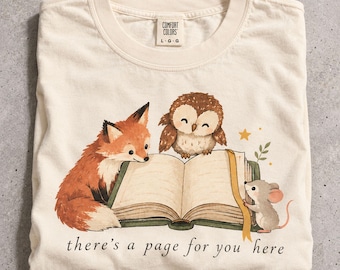 May include: An off-white t-shirt featuring a graphic of a fox, an owl, and a mouse reading an open book. The text "there's a page for you here" is printed below the illustration. The shirt has a Comfort Colors brand label at the inner neckline.