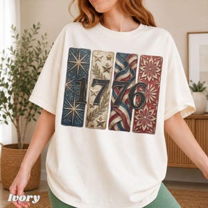 May include: Ivory-colored t-shirt featuring a patriotic design with the number "1776" in black. The design incorporates stars, stripes, and floral patterns in red, white, and blue, evoking a vintage aesthetic. The shirt has short sleeves and a relaxed fit.