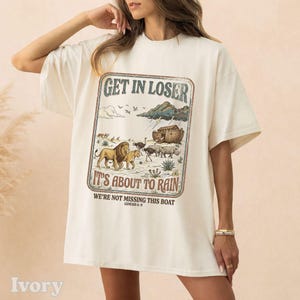 Op de afbeelding: Ivoor kleurig T-shirt met een grafische afbeelding met de tekst "GET IN LOSER IT'S ABOUT TO RAIN" en een illustratie van dieren en de ark. De tekst "WE'RE NOT MISSING THIS BOAT GENESIS 6-9" is ook opgenomen.
