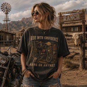 May include: A charcoal gray short-sleeve t-shirt featuring a graphic print of a skeleton riding a motorcycle in a desert landscape. The text RIDE WITH CONFIDENCE and HANG ON ANYWAY is printed in a tan font above and below the central illustration.