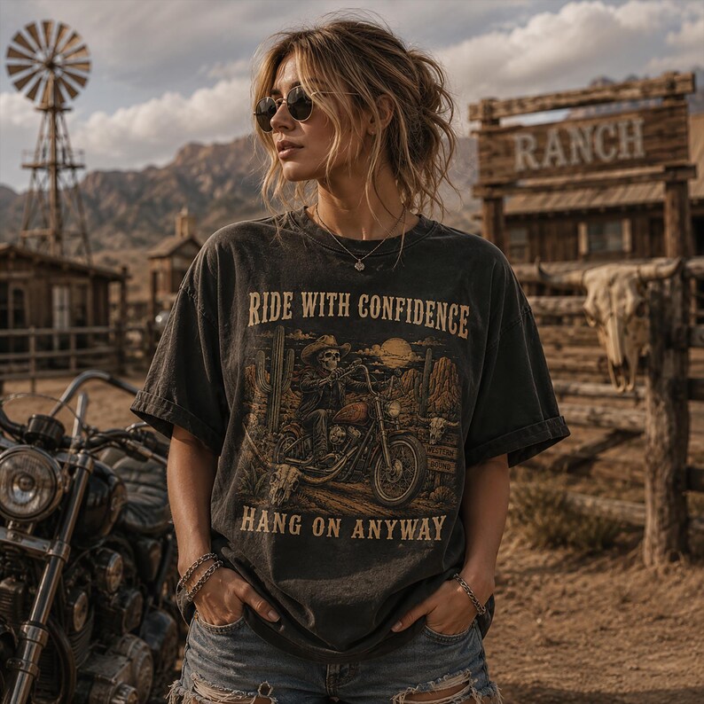 May include: A charcoal gray short-sleeve t-shirt featuring a graphic print of a skeleton riding a motorcycle in a desert landscape. The text RIDE WITH CONFIDENCE and HANG ON ANYWAY is printed in a tan font above and below the central illustration.