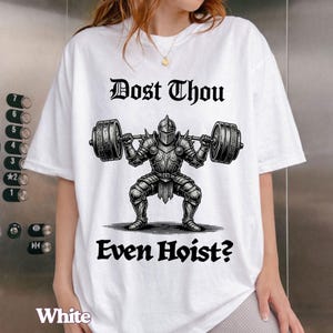 Op de afbeelding: Wit T-shirt met een zwarte afbeelding van een ridder die een halter optilt, en de tekst "Dost Thou Even Hoist?" in een gotisch lettertype. Het woord "White" staat onderaan het shirt.