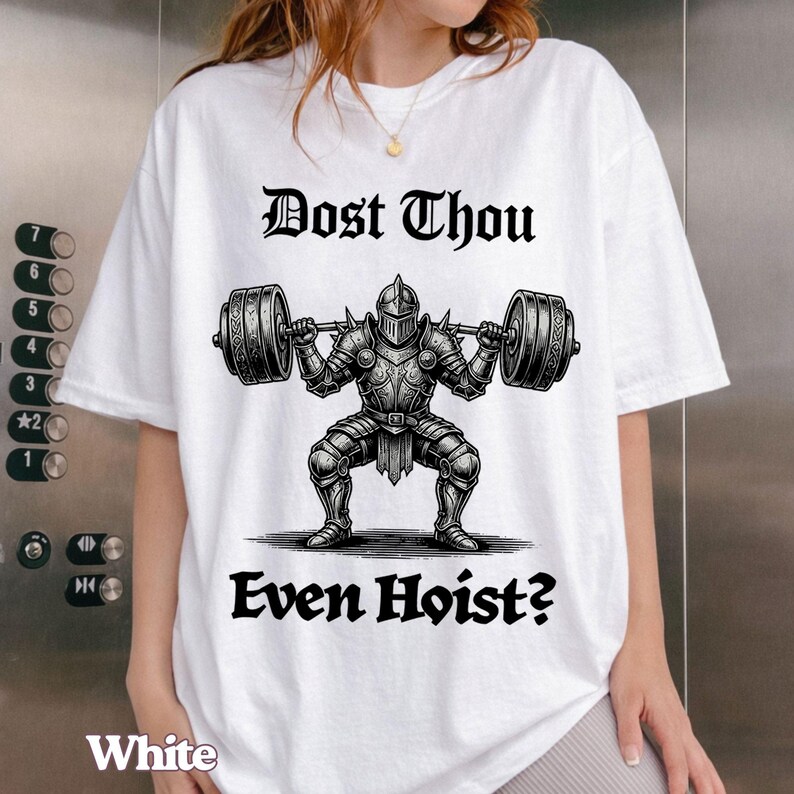Op de afbeelding: Wit T-shirt met een zwarte afbeelding van een ridder die een halter optilt, en de tekst "Dost Thou Even Hoist?" in een gotisch lettertype. Het woord "White" staat onderaan het shirt.