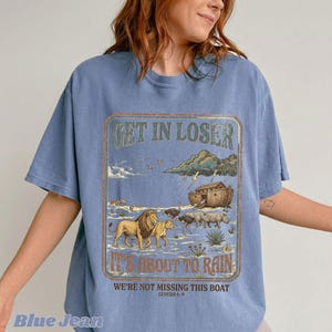 Op de afbeelding: Blauw T-shirt met ronde hals en grafisch ontwerp. Het ontwerp bevat de tekst "GET IN LOSER, IT'S ABOUT TO RAIN" met een illustratie van dieren, de ark en onweerswolken. De tekst "WE'RE NOT MISSING THIS BOAT" en "GENESIS 6-9" zijn ook opgenomen.