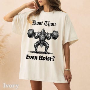 Op de afbeelding: Ivoor kleurig T-shirt met een zwarte afbeelding van een ridder die een halter optilt, met de tekst "Dost Thou Even Hoist?" erboven en eronder. Het shirt heeft korte mouwen en een relaxte pasvorm.