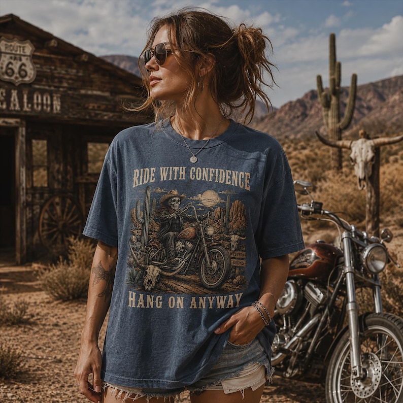 May include: A blue short-sleeve t-shirt featuring a graphic print of a skeleton wearing a cowboy hat and riding a motorcycle in a desert landscape. The text RIDE WITH CONFIDENCE and HANG ON ANYWAY is printed in a vintage font above and below the graphic.